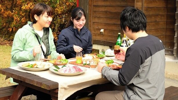 Trois personnes sont assises à une table en bois à l’extérieur, sur des bancs de pique-nique, avec en arrière-plan un paysage automnal et un bâtiment rustique.