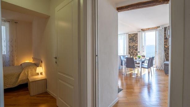 A bedroom with a bed, bedside table, and lamp. There is a dining area with a table and chairs. The room has wooden flooring and a stone wall. - Apartment in Lenno  Lake Como (Lenno)