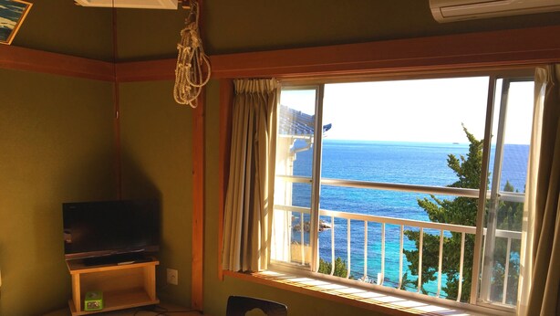 A room with a large window overlooking the sea, a television on a stand, and a futon bed. - Kiemonso (Shimoda)