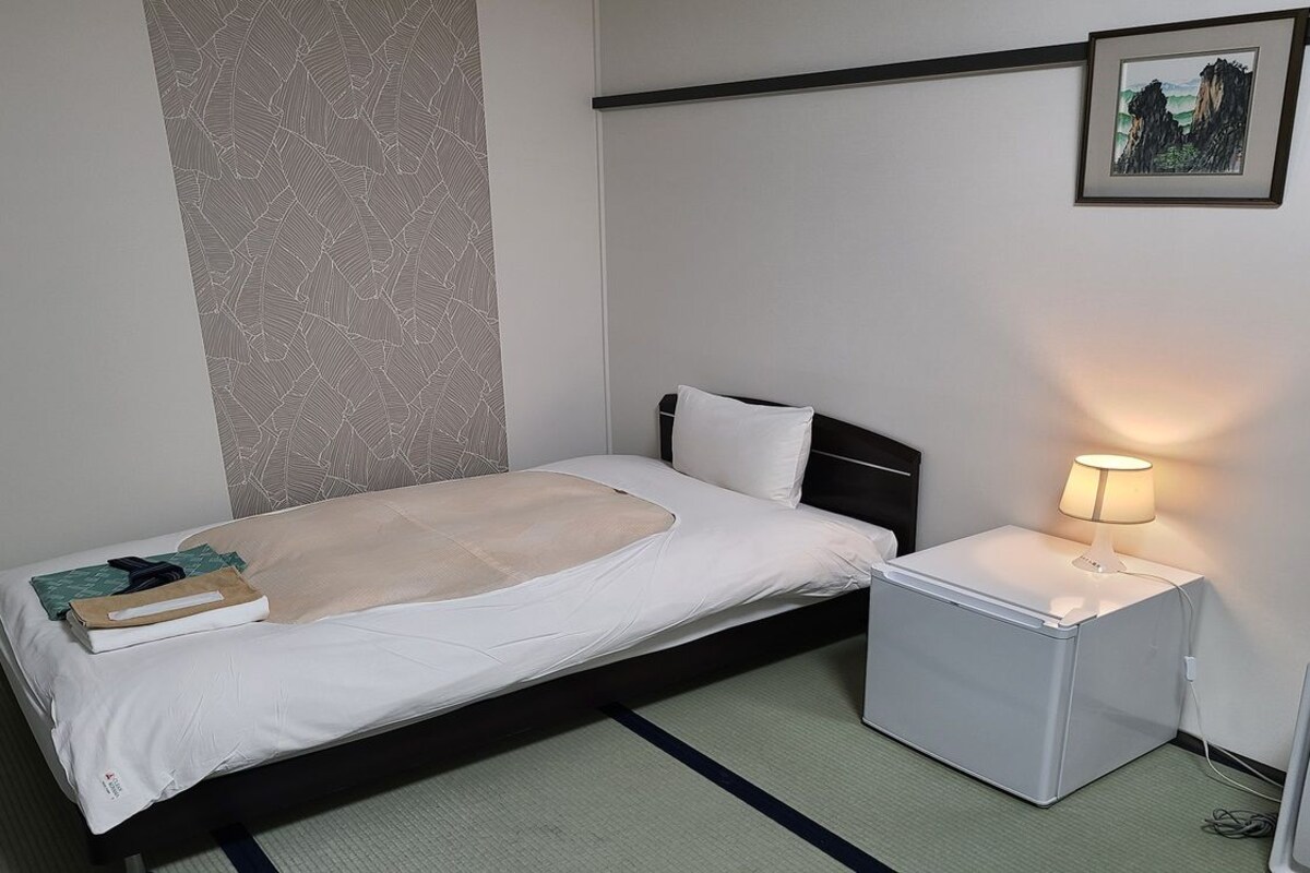Un lit simple avec un matelas blanc, une table de chevet avec une lampe et une petite armoire blanche.