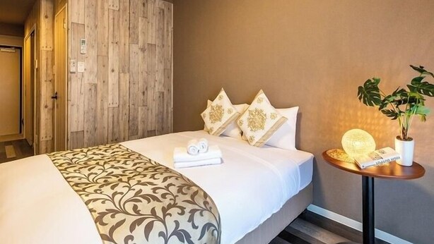 A hotel room with a bed, a wooden wardrobe, a small round table with a lamp, and a potted plant. - Mr. Kinjo in Mihara (Naha)