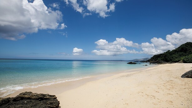 A beach with clear blue water, a sandy shore, and scattered clouds in the sky. - Akizero Onnason (Onna)