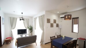 A modern living room with a flat-screen TV, a sofa, a dining table with chairs, and a large window with curtains. - Akizero Onnason (Onna)