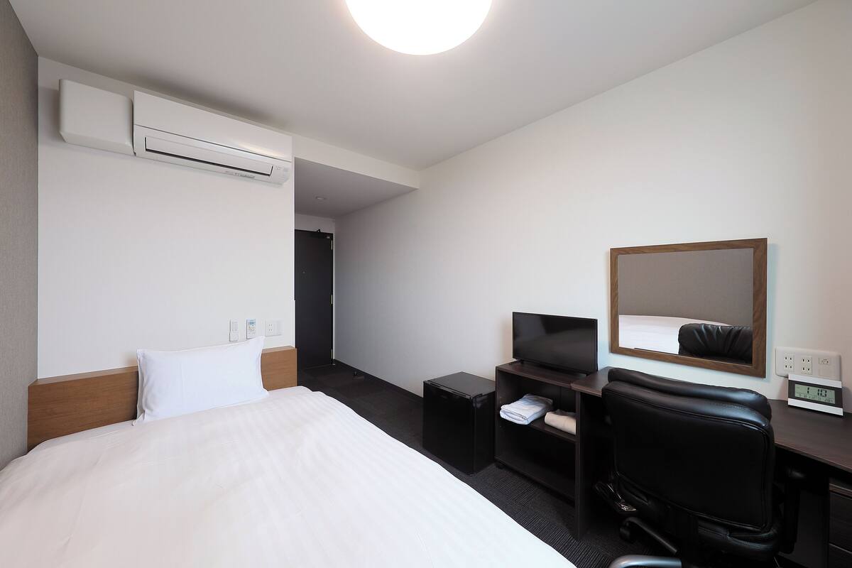 A hotel room with a bed, a desk with a chair, a television, a small table, a mirror, and an air conditioning unit.