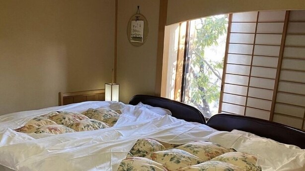 A neatly made bed with floral pillows, a bedside lamp, and a view of trees through shoji screens.