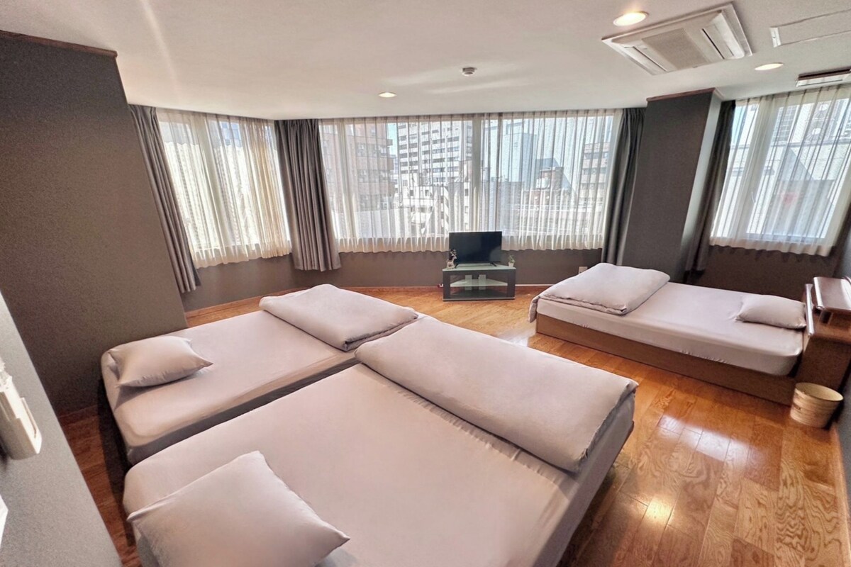 A modern hotel room with a large bed, a smaller sofa, a TV, and large windows with curtains.