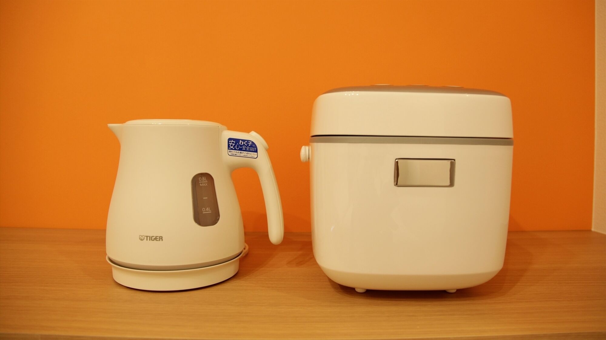 Two kitchen appliances, a white electric kettle and a white rice cooker, on a wooden surface against an orange background.