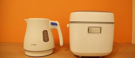 Two kitchen appliances, a white electric kettle and a white rice cooker, on a wooden surface against an orange background.