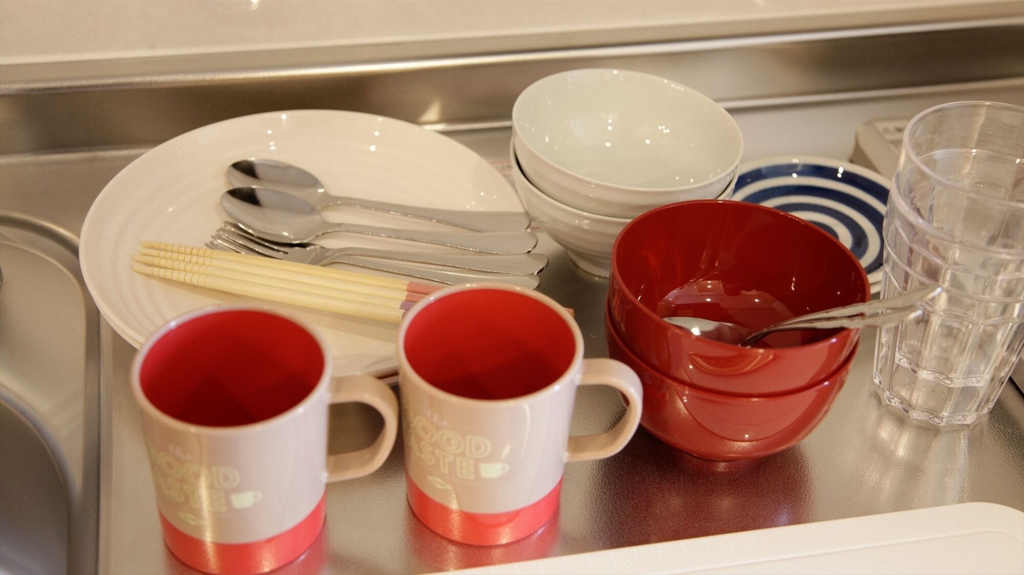 A kitchen counter with various utensils and dishes, including a plate with chopsticks, a red mug, and a clear glass.