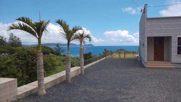 A coastal property with a gravel driveway, palm trees, and a view of the ocean. - Sol E Mar Amami Oshima (Tatsugo)
