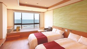 A hotel room with two beds, a large window with a view of the sea, a small seating area, and a textured wall.