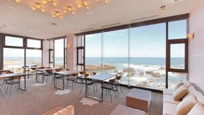 A modern dining area with large windows overlooking the ocean, featuring tables and chairs arranged for guests.