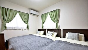 A bedroom with a large bed, two windows with green curtains, a white air conditioner, and a wooden nightstand.