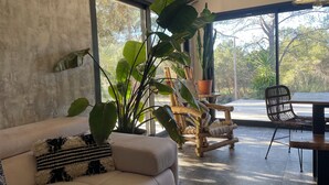 A modern living room with a beige sofa, a wooden rocking chair, and a dining table with chairs. Large windows provide a view of the outdoors. - Villa Luxury Lagoon - Aix en Provence (Ventabren)