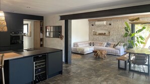 A modern kitchen with a built-in island, a dining area with a bench and chairs, and a living room with a sofa and a large plant. - Villa Luxury Lagoon - Aix en Provence (Ventabren)