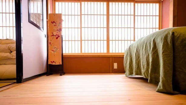 A room with a wooden floor, a bed with a green cover, a window with vertical blinds, and a decorative item with floral patterns. - Fujisan Kingyo (Fujikawaguchiko)
