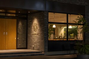 Entrada moderna de un edificio con puerta de madera, un logotipo iluminado grande y un área de estar con plantas visibles a través de las ventanas.