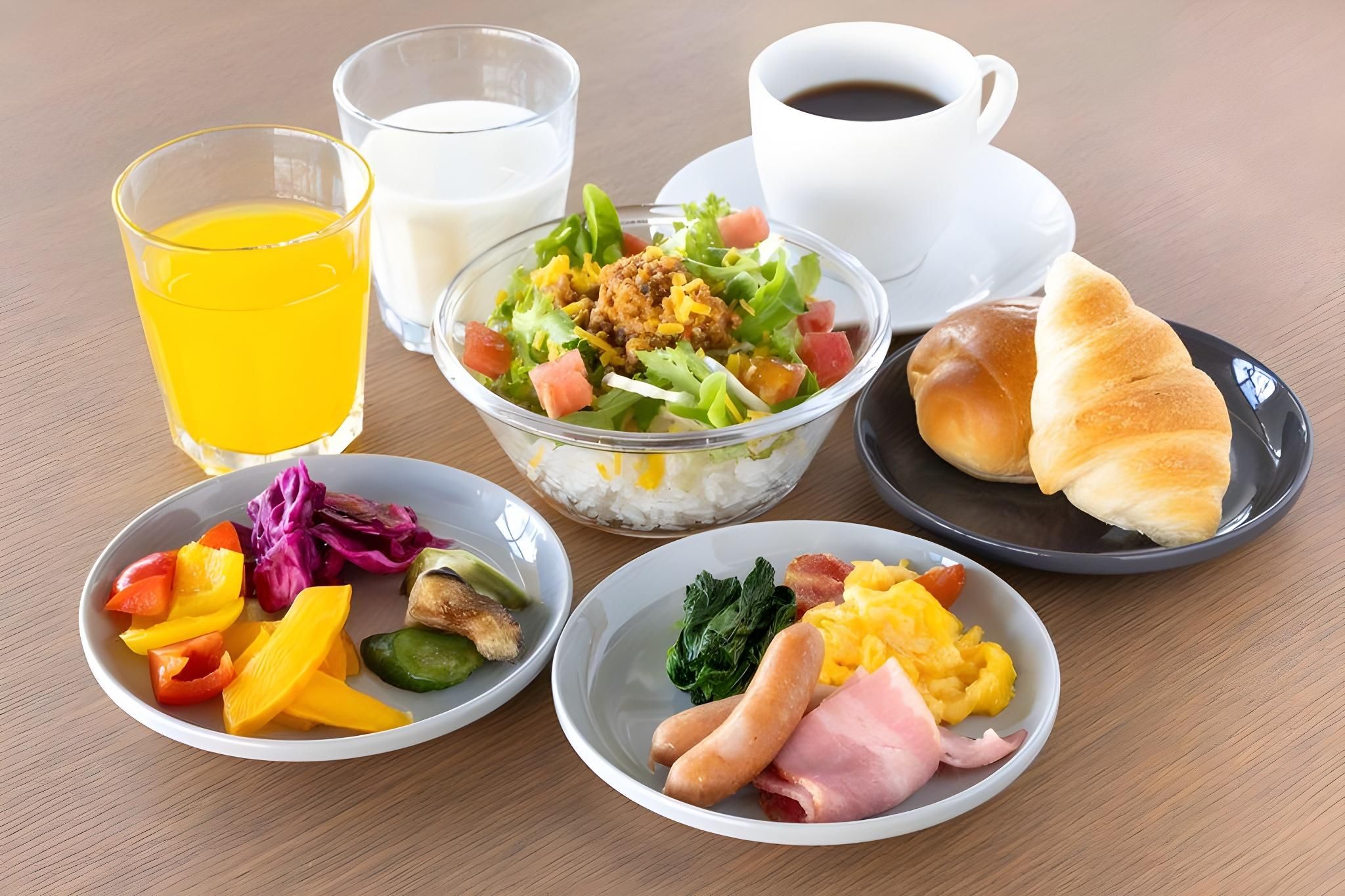 Une table où sont disposés divers aliments pour le petit-déjeuner, notamment un saladier de salade, une assiette d’œufs, une assiette de jambon, un verre de jus d’orange, une tasse de café et un verre de lait.