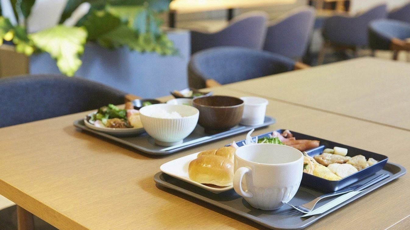 Une table à manger avec des plateaux contenant de la nourriture et une tasse blanche, placée dans une pièce où se trouvent des chaises et une plante en arrière-plan.