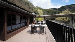 Um edifício tradicional japonês com telhado de palha e portas deslizantes, com vista para um rio e uma ponte.