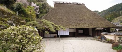 Un bâtiment japonais traditionnel, doté d’un toit de chaume, entouré d’une cour de gravier et d’une végétation luxuriante.