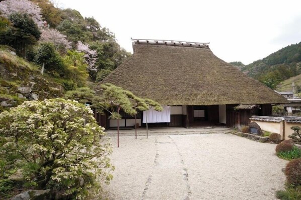 Un edificio japonés tradicional con techo de paja, rodeado por un patio de grava y abundante vegetación.