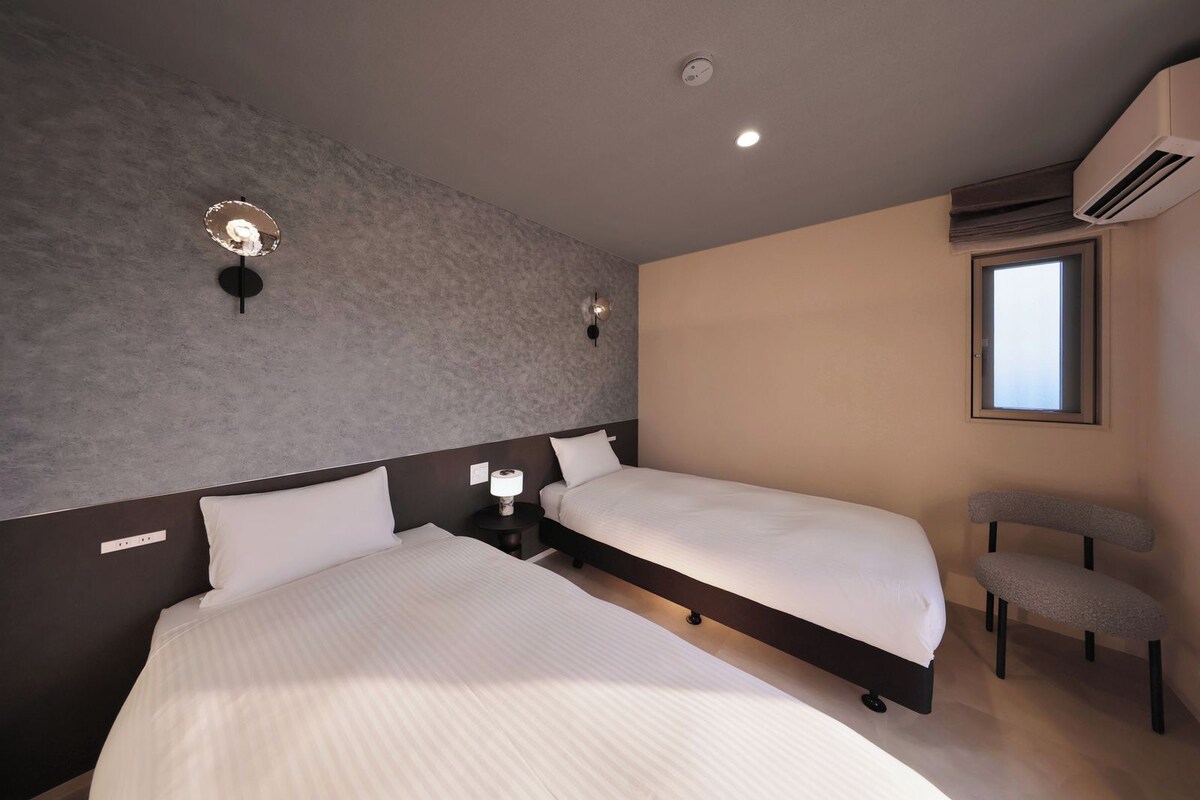 Quarto de hotel moderno com duas camas, uma mesinha, uma cadeira, uma janela e um ar condicionado.