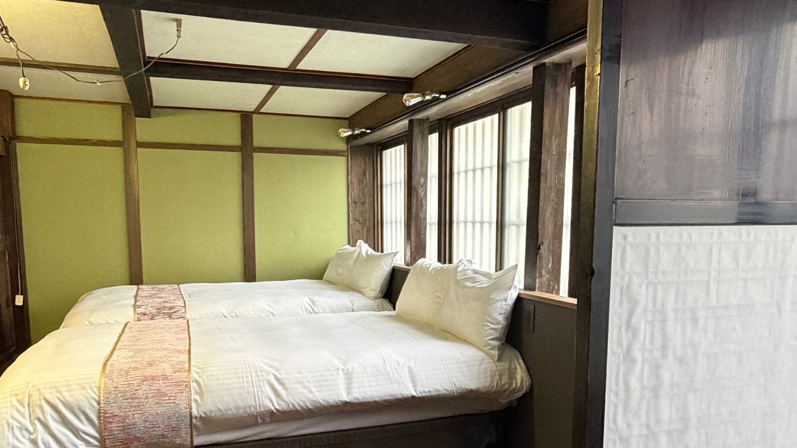 A room with two beds, wooden beams, a window with blinds, and a green wall.