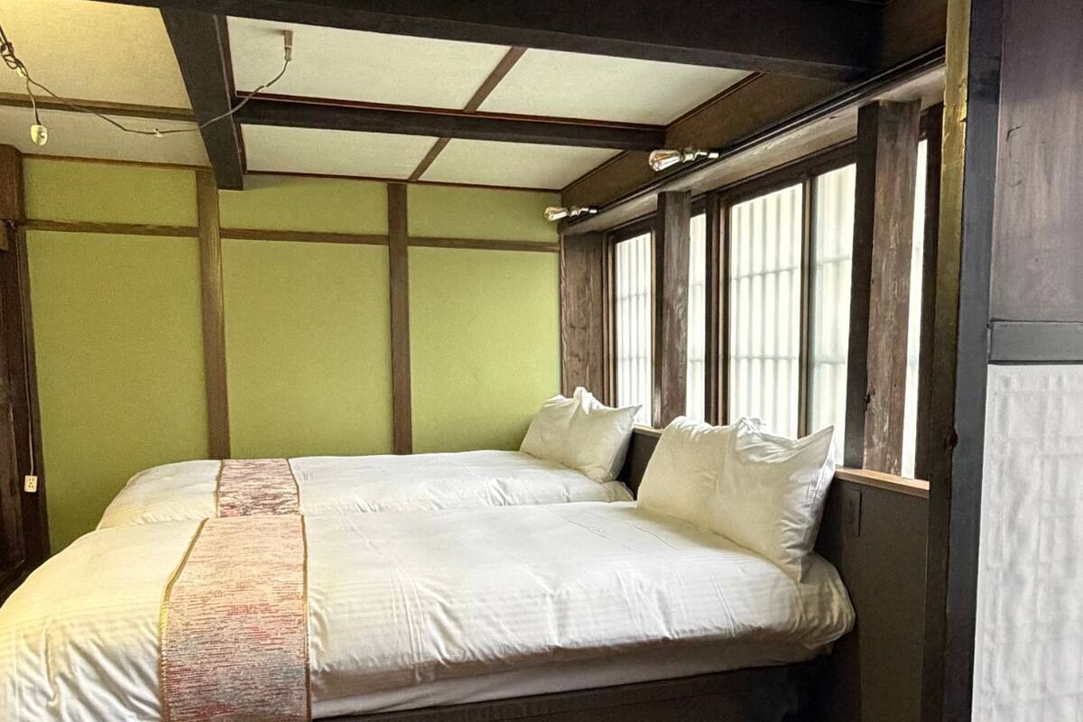 Een kamer met twee bedden, houten balken, een raam met zonwering en een groene wand.