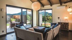 A spacious living area with a sofa set, a dining table, and a lamp. Large sliding glass doors lead to a balcony with a view of mountains. - Toki Resort Yufuin Villa (Yufu)