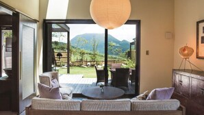 A room with a sofa, a table, and a view of a mountainous landscape through large glass doors. - Toki Resort Yufuin Villa (Yufu)