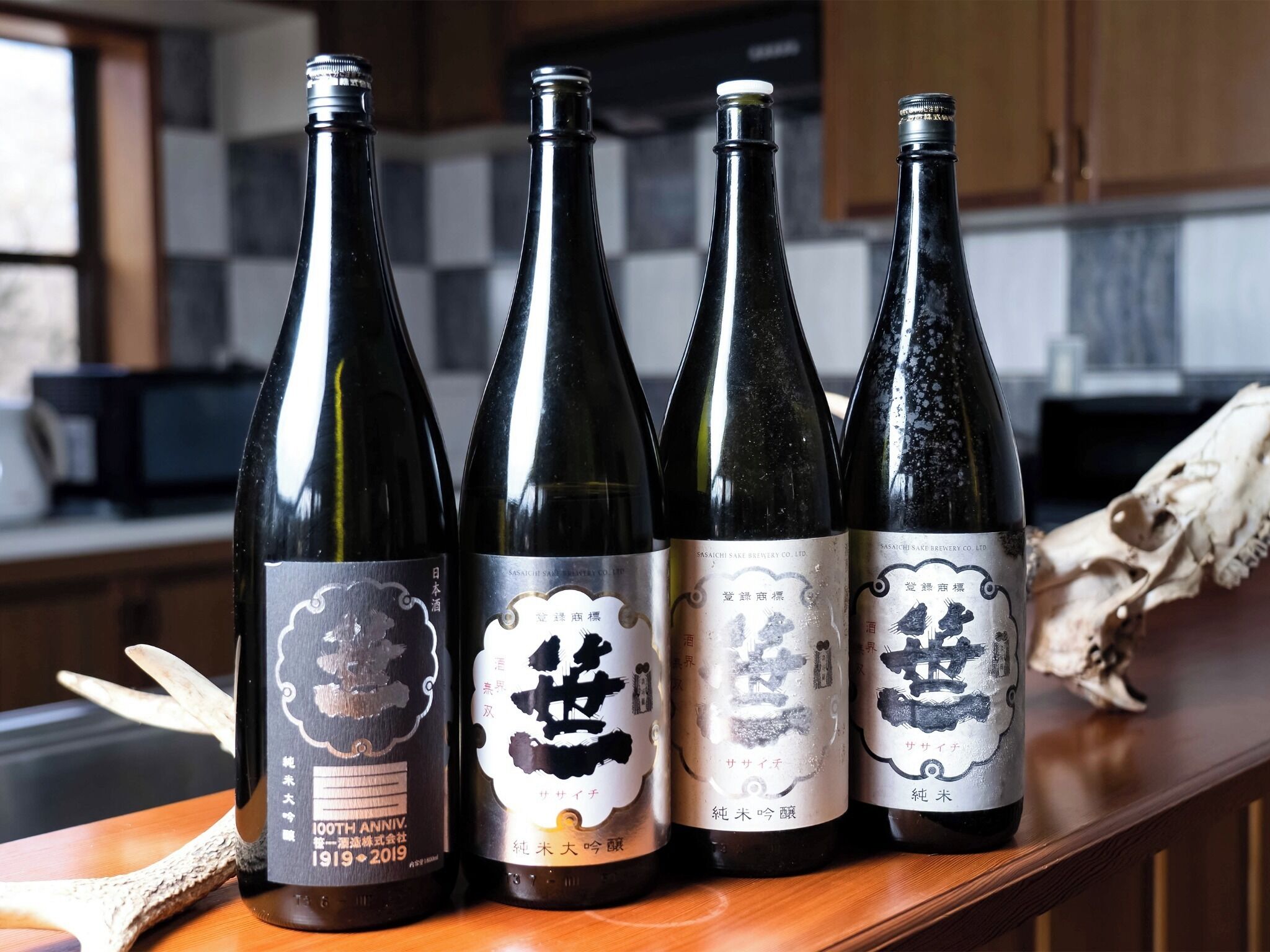 Four bottles of sake on a wooden surface with a kitchen in the background.