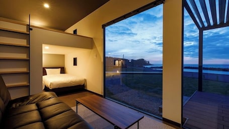 A modern bedroom with a bed, a sofa, and a large window offering a view of the sea and a distant island.