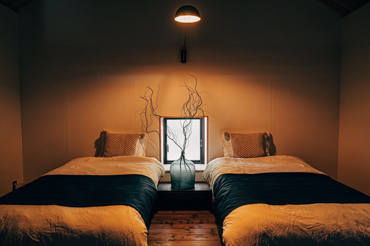 A room with two beds, a window, and a vase on the windowsill.