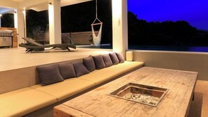 En modern utomhusplats med sittmöbler, bestående av ett träbord och en inbyggd eldstad, en soffa med kuddar och en hängstol.