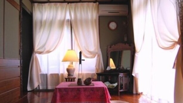 A room with a wooden floor, a table with a red tablecloth, a lamp, a mirror, and a clock on the wall. - Milk-ya Women-only Guest House (Yomitan)