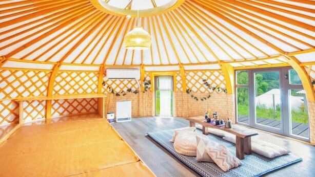Eine Jurte mit einem Bett in der Mitte, einem kleinen Tisch mit Gegenständen und Blick auf grüne Landschaft.