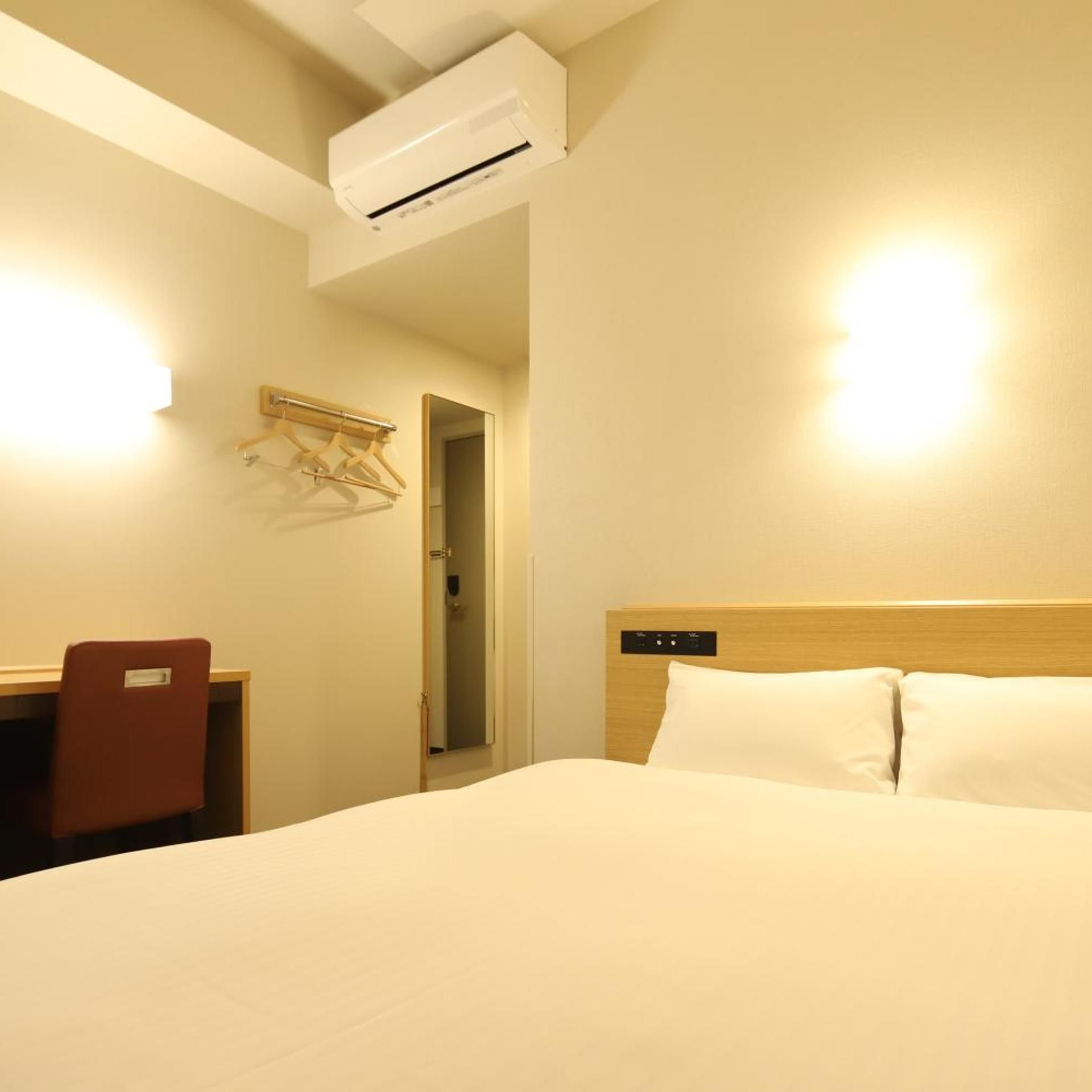 A hotel room with a bed, a desk, a chair, an air conditioning unit, and a mirror.
