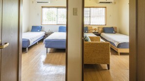 A room with two beds, a wicker chair, and a window with blinds. - Panorama (Yomitan)