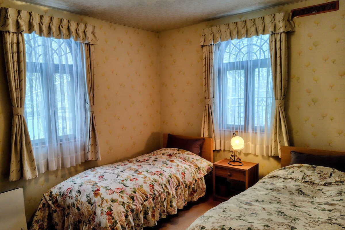 Um quarto com duas camas de solteiro, uma mesa de cabeceira com abajur e um guarda-roupa.