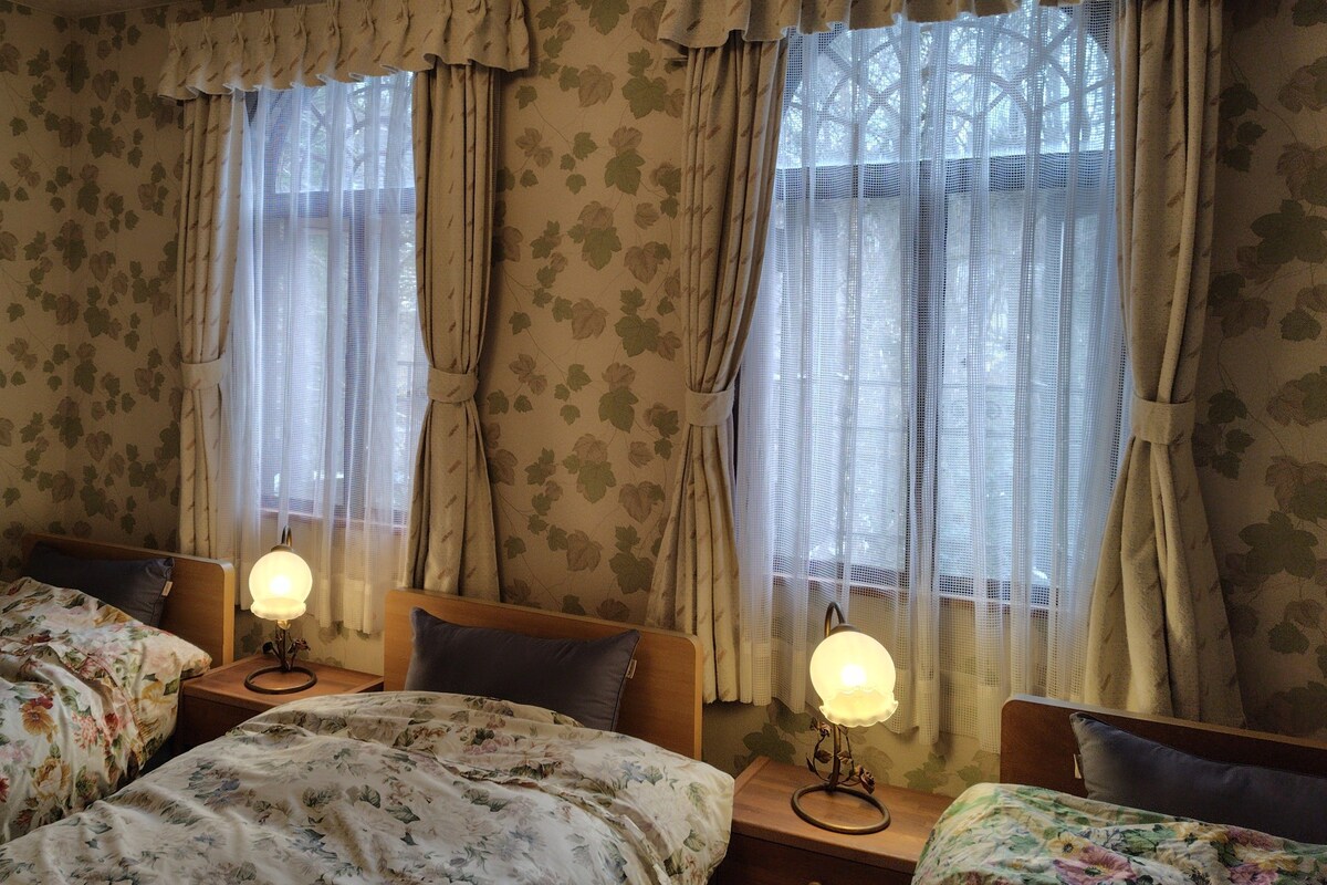 Quarto com duas camas de solteiro, roupa de cama estampada com flores e mesa de cabeceira com abajur entre as camas.