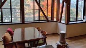 Un coin repas avec une table et des chaises en bois, un poêle à bois et de grandes fenêtres offrant une vue sur le feuillage automnal.