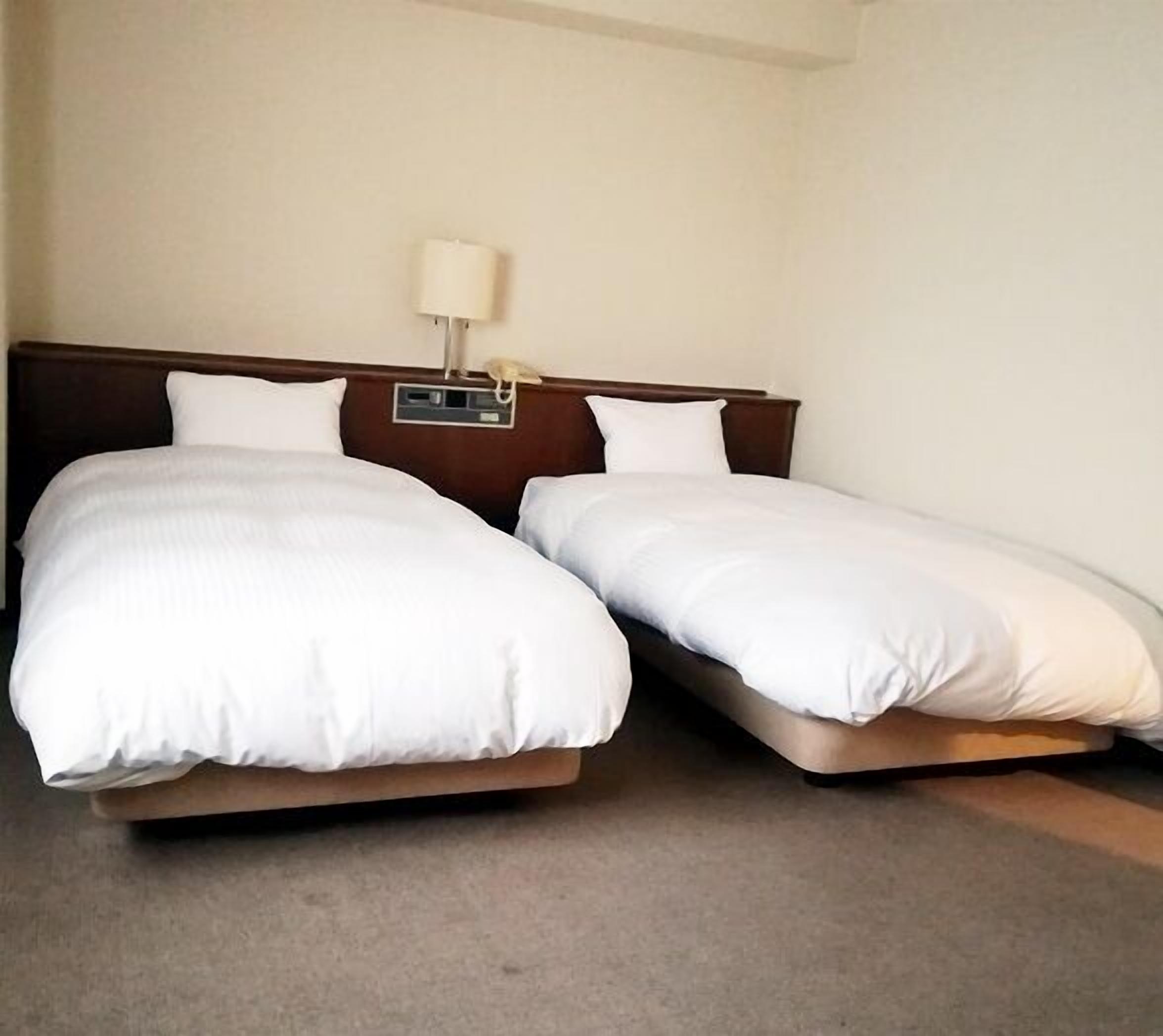 A hotel room with two single beds, a nightstand with a lamp, and a plain wall.