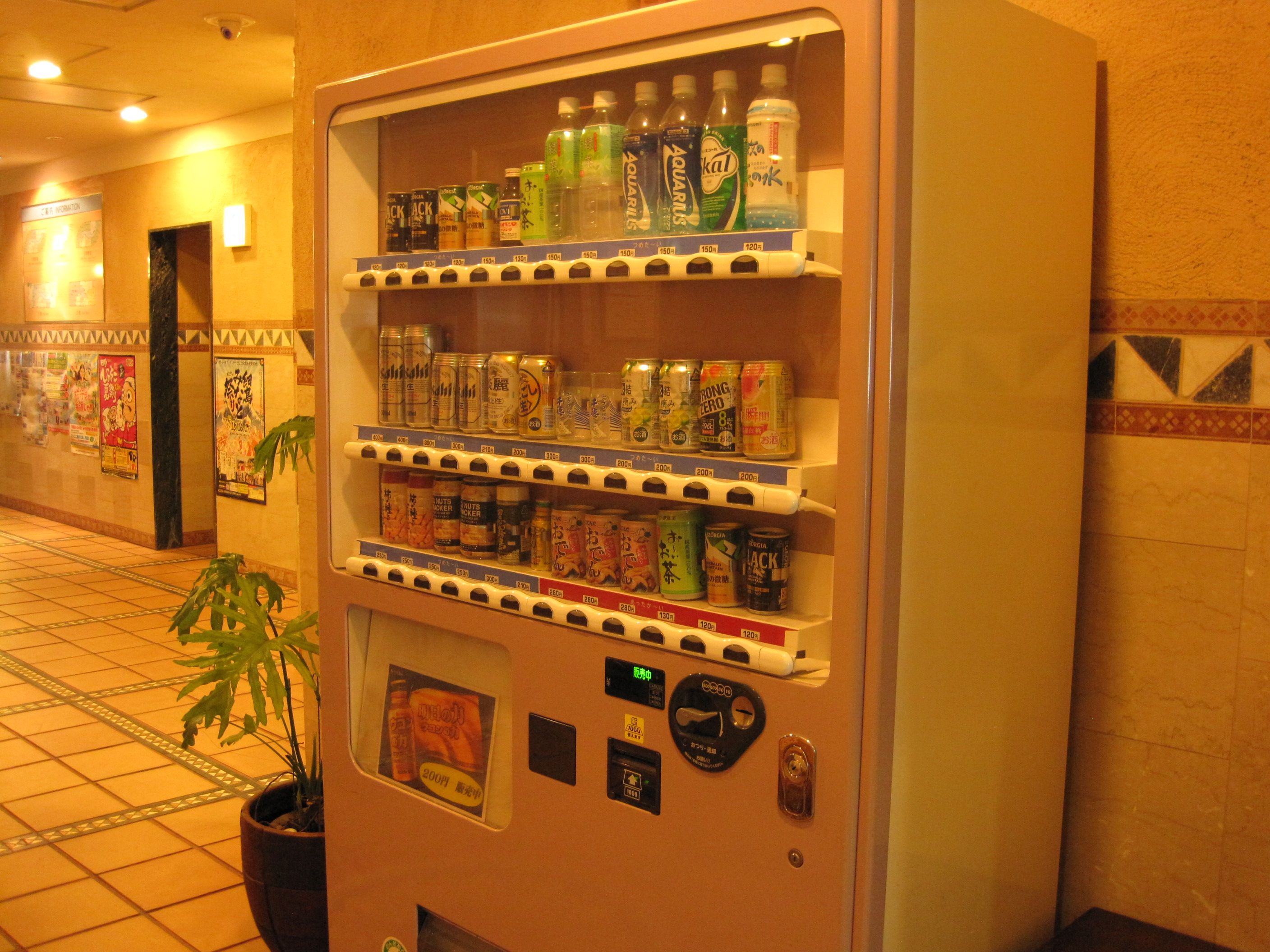 A vending machine with various beverages and snacks, located in a hallway with tiled flooring and a potted plant.