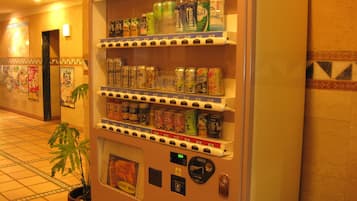 A vending machine with various beverages and snacks, located in a hallway with tiled flooring and a potted plant.