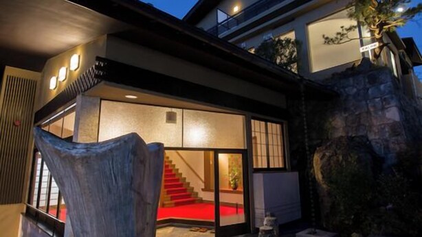 A modern house with a large wooden sculpture in the foreground, a red carpet leading to a staircase, and a stone wall on the right. - Yado Atagoya (Fukushima)