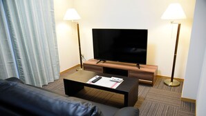 A hotel room with a flat-screen TV on a wooden stand, two lamps, a black leather sofa, and a coffee table with a remote control and a menu. - Nemuru Kagoshima Chuo Eki (Kagoshima)