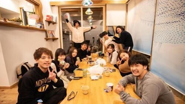 Ryhmä ihmisiä istuu puisen pöydän ympärillä huoneessa, jossa on puulattia, seinällä valkotaulu ja taustalla ovi.