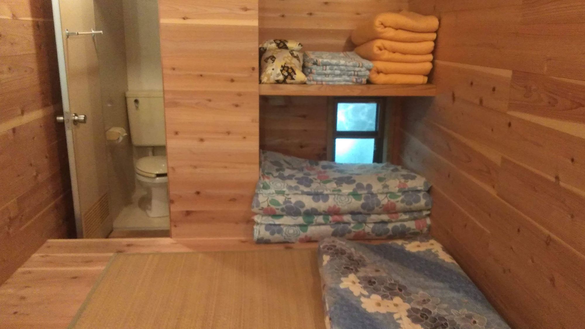 A compact, wooden cabin-style room with a small bed, a toilet, and a window.