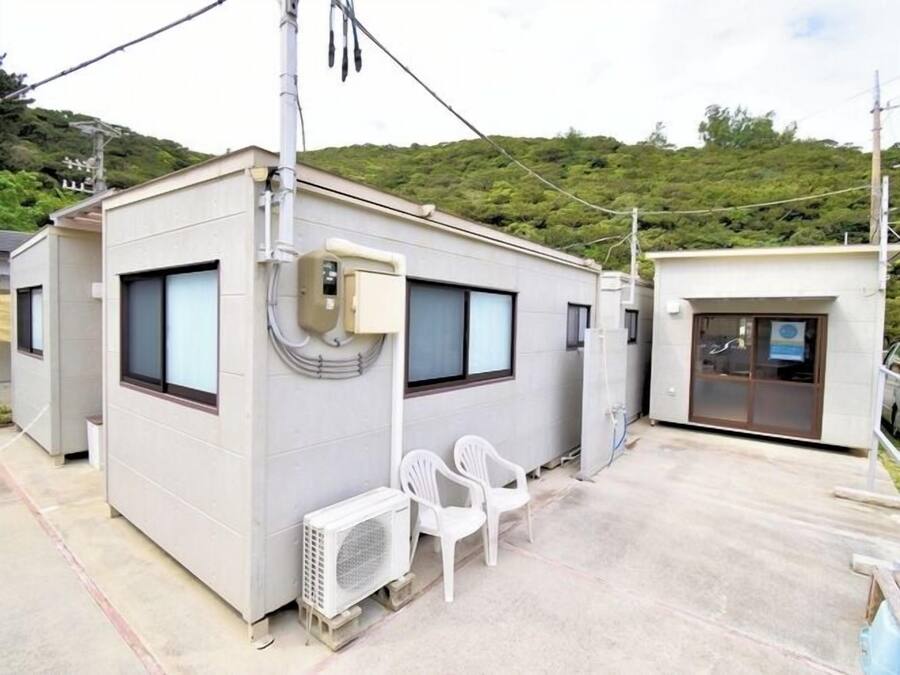 A small, single-story building with a flat roof, a window, and a door. There are two white plastic chairs outside and a white air conditioning unit on the ground.